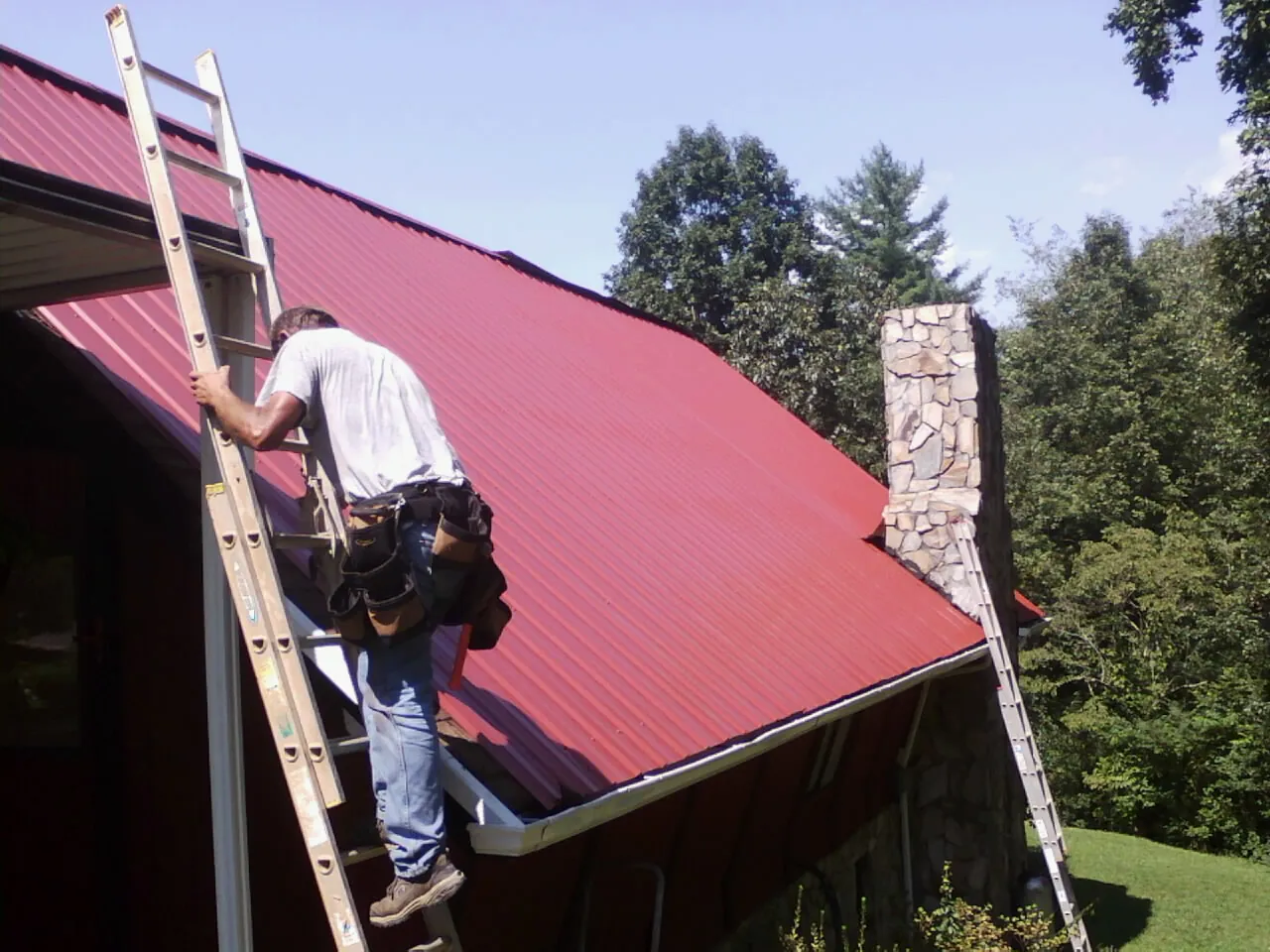 Roofer on a ladder beside a red metal roof and stone chimney — Soffit & Fascia Repair by Roofing Brothers in Yarmouth